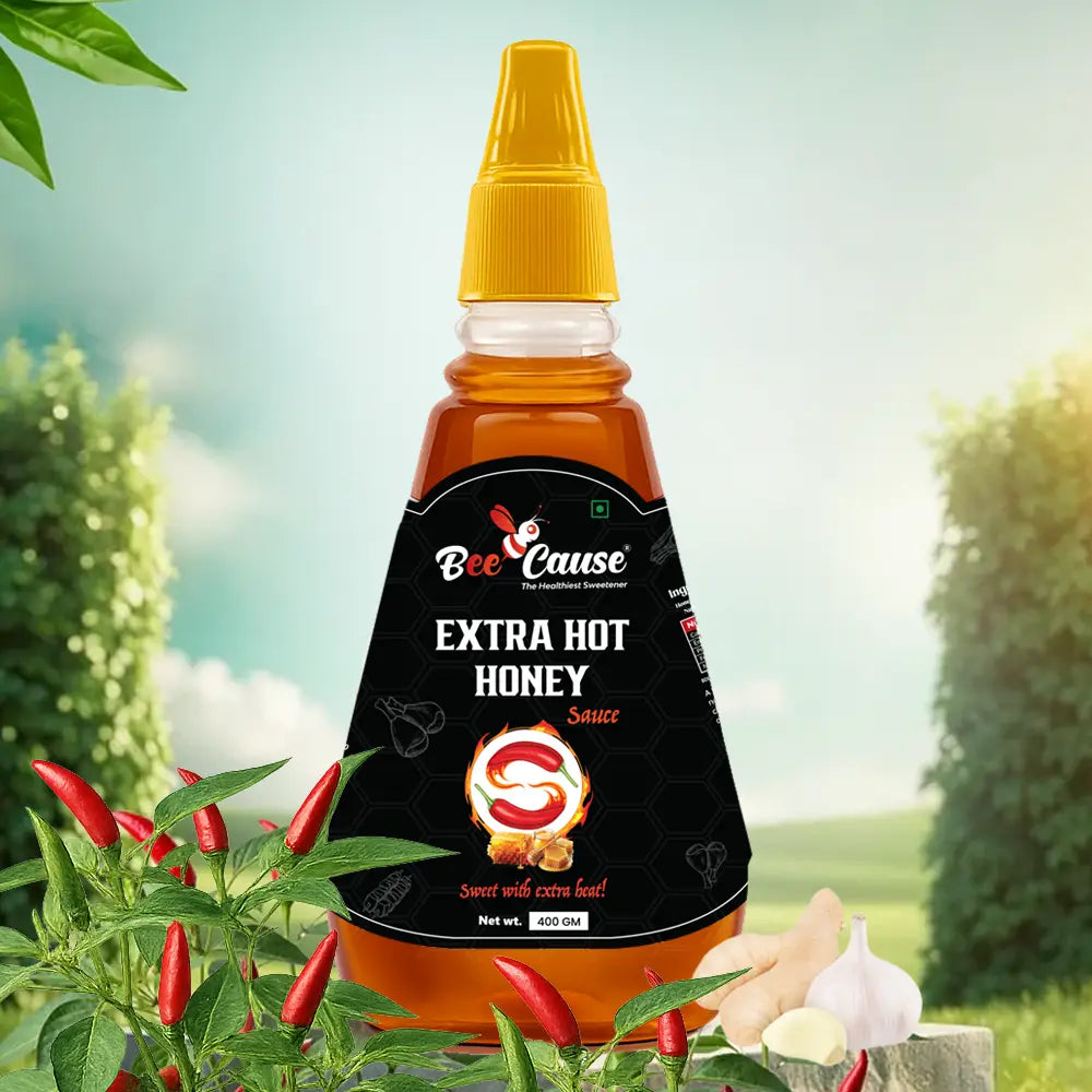 A bottle of Bee Cause Extra Hot Honey Sauce featuring a black label with a red chili graphic and the text "Sweet with extra heat!". It is displayed outdoors on a stone surface, surrounded by fresh red chili peppers growing on a plant, raw ginger, and garlic cloves, set against a bright, sunny sky and green fields.