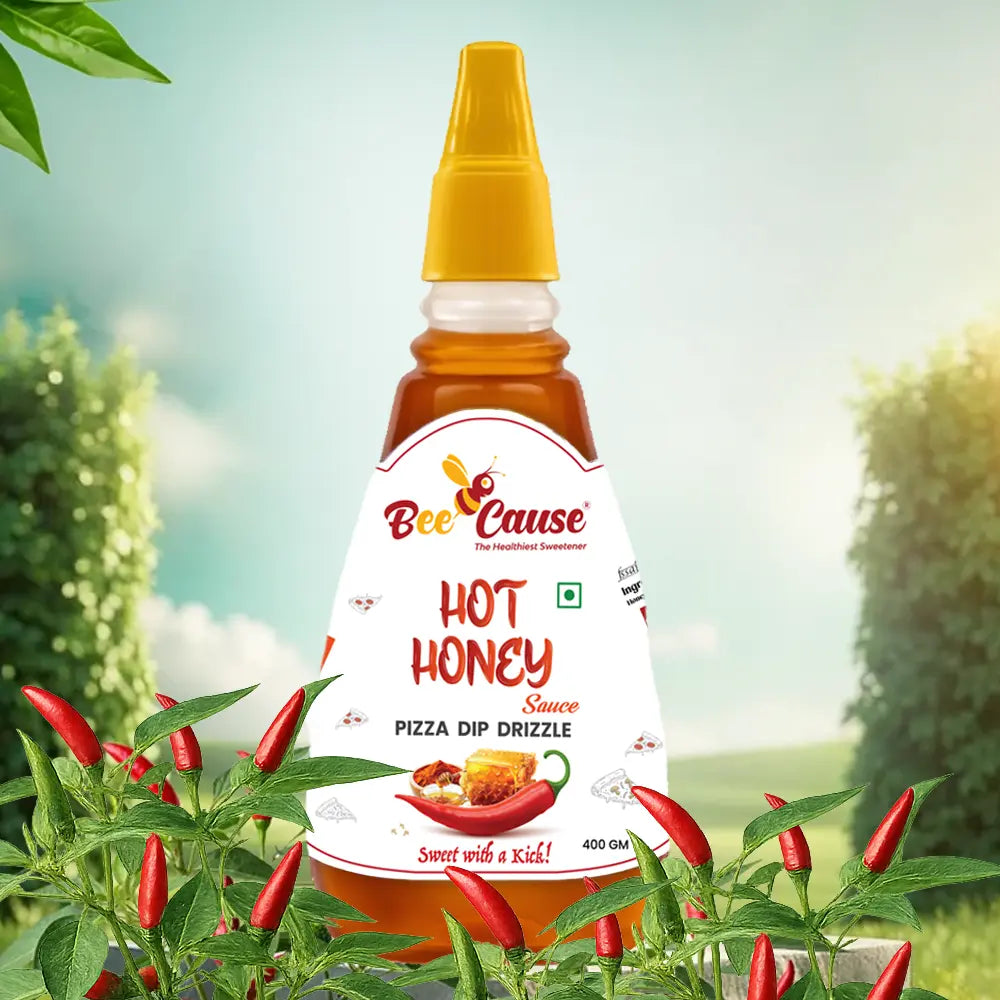 A bottle of Bee Cause Hot Honey Sauce with a white label that reads "Pizza Dip Drizzle" and "Sweet with a Kick!". The bottle is placed outdoors among vibrant red chili plants with green leaves, with a bright, sunny meadow and blurred trees in the background.