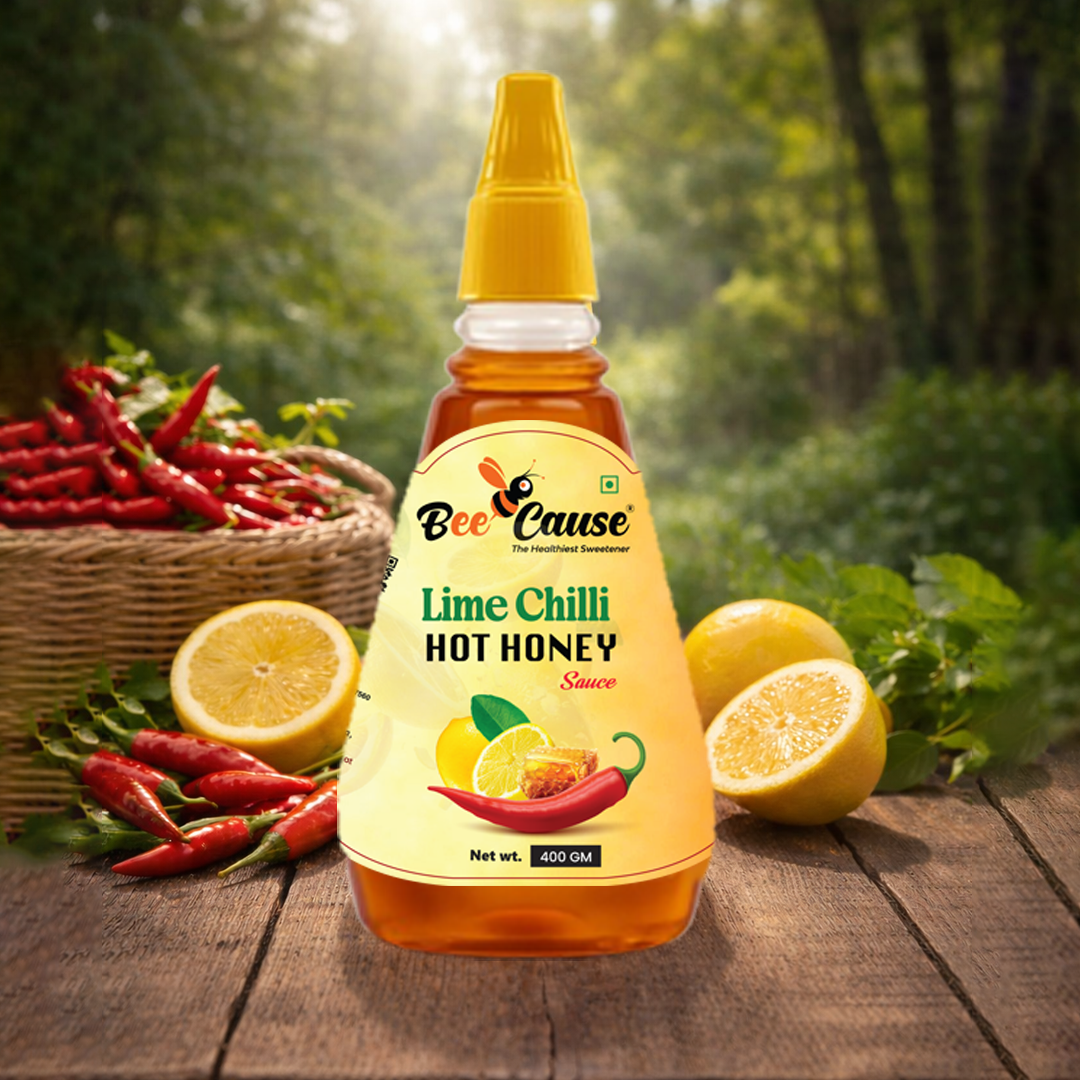 A squeeze bottle of Bee Cause Lime Chilli Hot Honey Sauce on a wooden table, surrounded by fresh halved lemons and a woven basket overflowing with bright red chilies against a green, leafy background.