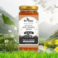 A single jar of Bee Cause Multiflora Raw Golden Honey sitting on a grassy bank next to a small stream, surrounded by white and yellow wildflowers.