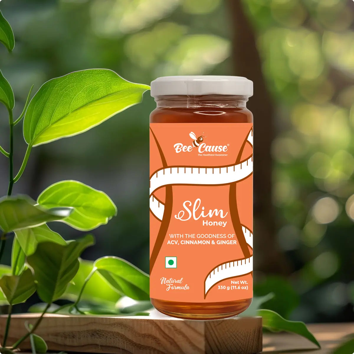 A glass jar of Bee Cause Slim Honey with a label featuring a measuring tape design. The label highlights natural ingredients including Apple Cider Vinegar, Cinnamon, and Ginger, set against a blurred background of vibrant green leaves.
