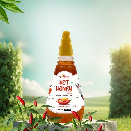 A scenic outdoor product display of Bee Cause Hot Honey Sauce, labeled as a "Pizza Dip Drizzle." The squeeze bottle is set amidst chili plants and features pizza graphics with the tagline "Sweet with a Kick".
