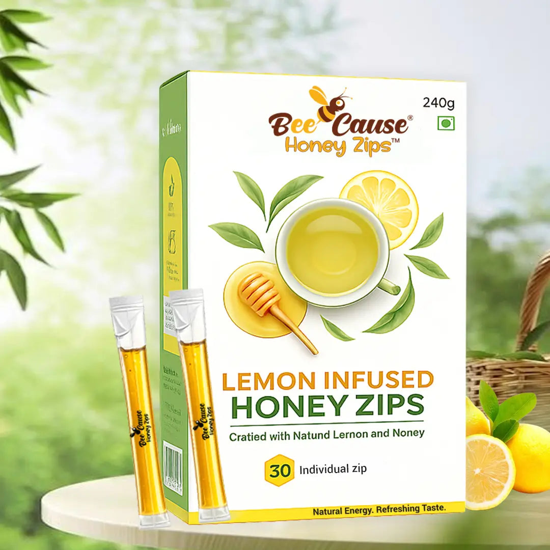 A lifestyle product shot of the Bee Cause Lemon Infused Honey Zips box standing on a round table in a sunny outdoor setting with green foliage. Two individual honey sachets stand upright next to the 240g box, with fresh lemons in a basket in the background.