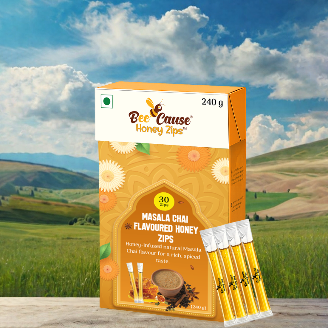 A 240-gram box of Bee Cause Masala Chai Flavoured Honey Zips and four individual honey sticks placed on a wooden surface against a picturesque background of green hills and a partly cloudy blue sky.