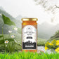 A scenic product display of Bee Cause Multiflora Raw Golden Honey placed on a wooden stump near a flowing stream, surrounded by lush green grass and mountains.