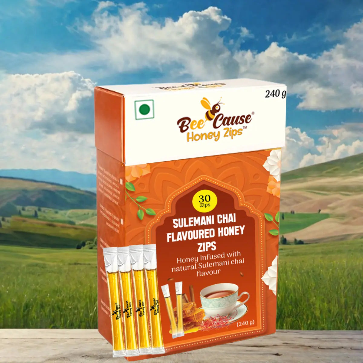 A 240g box of Bee Cause Sulemani Chai Flavoured Honey Zips and four individual honey sticks displayed on a wooden surface against a scenic background of rolling green hills and a cloudy blue sky.