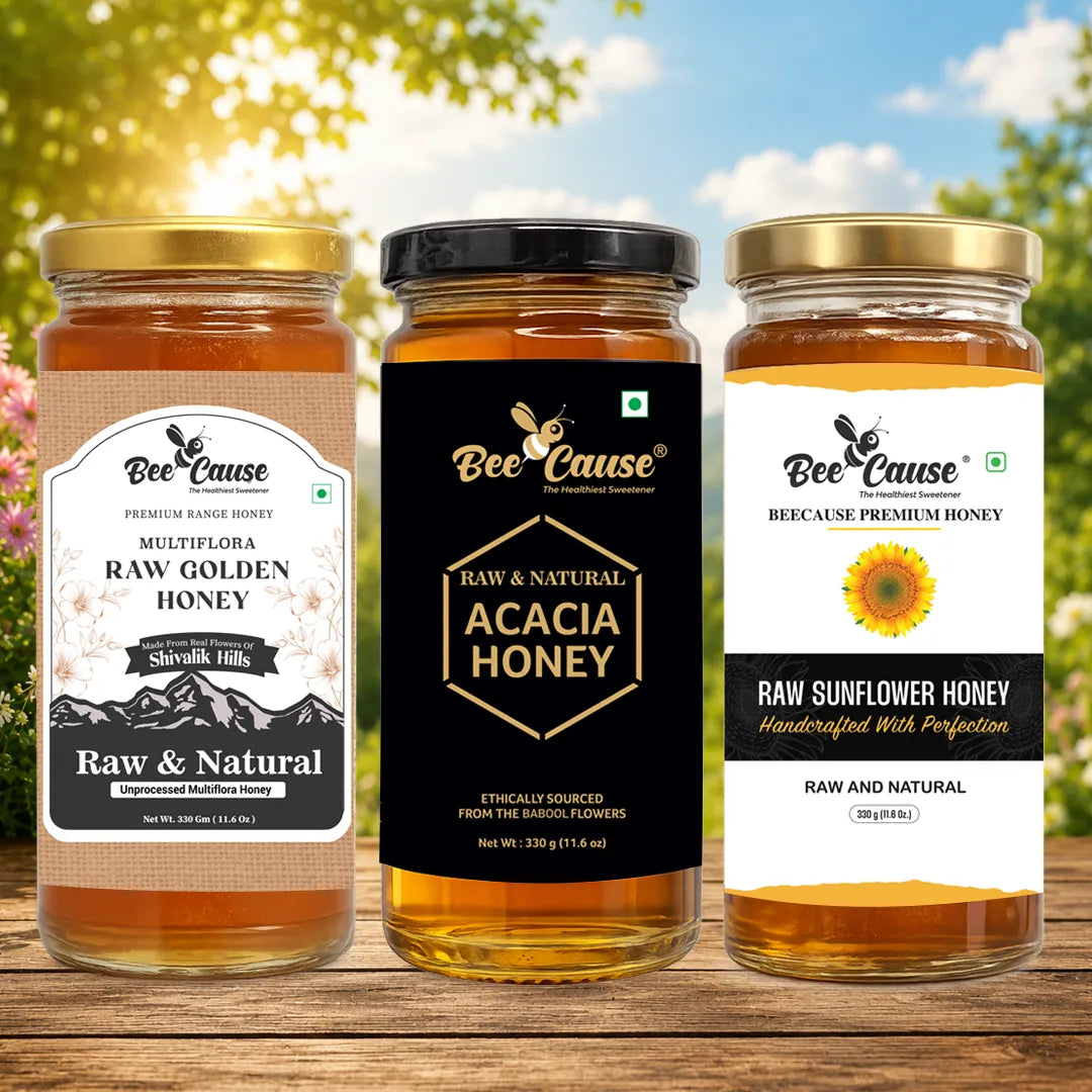 Three glass jars of Bee Cause honey arranged on a wooden surface outdoors, with a blurred sky and greenery background. The left jar is Raw Golden Honey, the center jar is Acacia Honey, and the right jar is Raw Sunflower Honey.