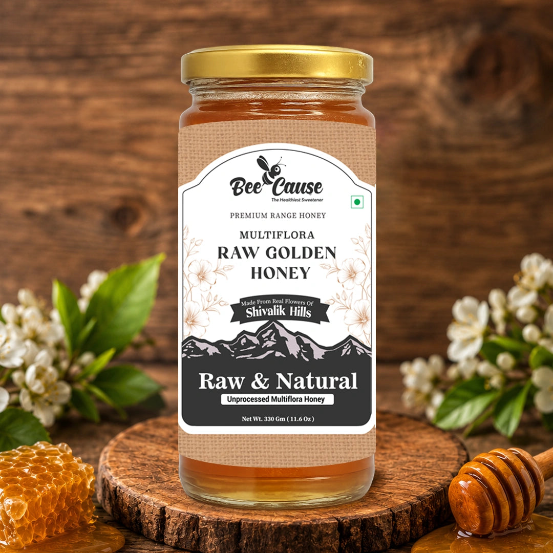 A glass jar of Bee Cause Raw Golden Honey with a gold lid and beige label, displayed on a rustic wooden slice with honeycomb, white blossoms, and a wooden honey dipper against a dark wood background.