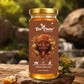 A jar of Bee Cause Sulai Honey displayed on a rustic wooden surface with a blurred natural background of rocks and greenery. The jar has a gold lid and a brown label with honeycomb and bee illustrations.
