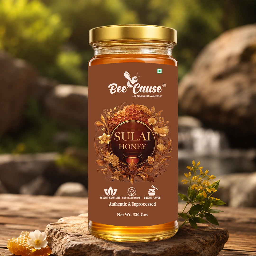A jar of Bee Cause Sulai Honey displayed on a rustic wooden surface with a blurred natural background of rocks and greenery. The jar has a gold lid and a brown label with honeycomb and bee illustrations.