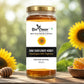 A glass jar of Bee Cause Raw Sunflower Honey with a gold lid and white label, set on a wooden surface with sunflowers and honeycomb in a blurred garden background.